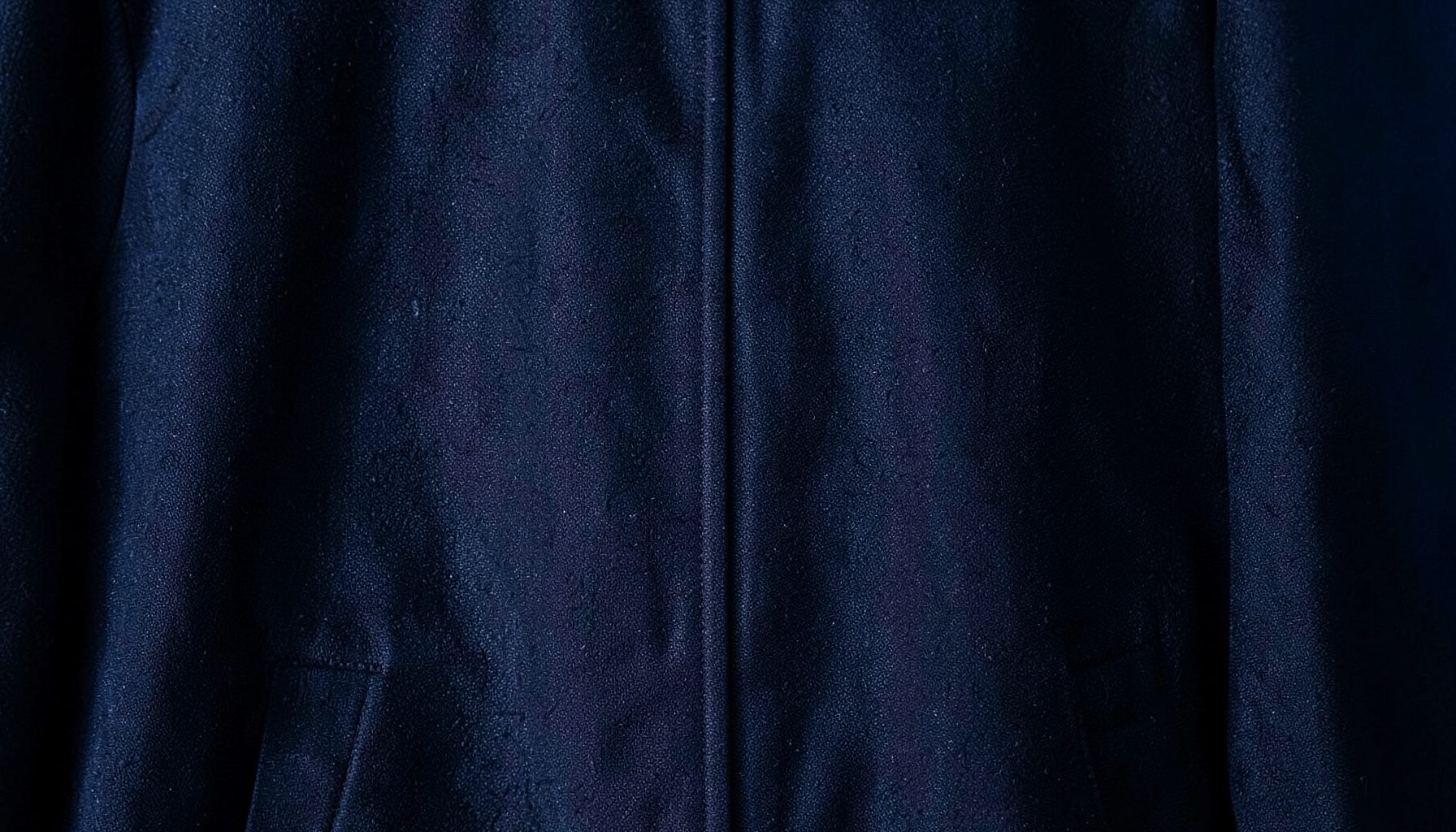 Dark atmospheric photograph of a textured jacket fabric with subtle sheen, deep navy and charcoal tones, moody and minimal